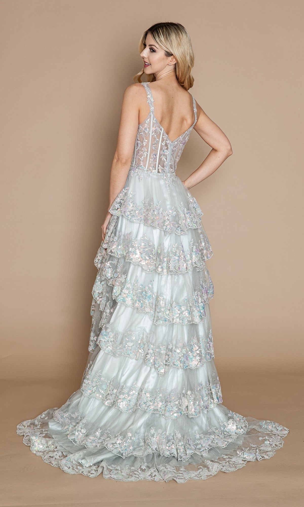 A woman faces away in the Sheer-Corset Long Ruffled Prom Dress (Poly USA 9410): a light blue, floor-length gown featuring floral embroidery, a sheer corset bodice, and delicate tiers, set against a plain beige background.